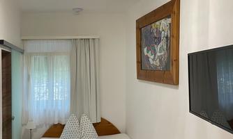A simple bedroom with a double bed, a television, and a table. The room has large windows and a calm, bright atmosphere.