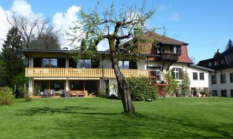 A large house with a balcony and a beautiful garden. In the background, there are trees and the sky is blue with a few clouds.