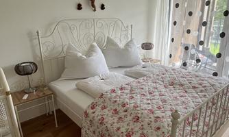 A cozy bedroom with a white iron bed and pink floral bedding. Bright curtains provide plenty of natural light.