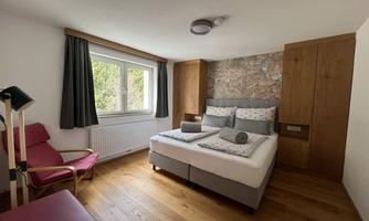A bright bedroom with a large bed and modern furniture. There is also a cozy armchair and wooden flooring.
