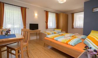 A cozy bedroom with two beds and colorful bed covers. The room has large windows with curtains and a small dining area with a table and chairs.