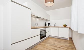 A modern kitchen with white cabinets and a wooden countertop. It is bright and minimally furnished.