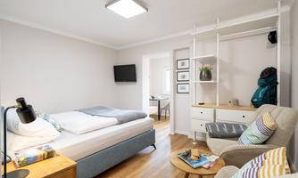 A cozy bedroom with a large bed, a television, and an armchair. The decor is modern and bright, perfect for relaxation.