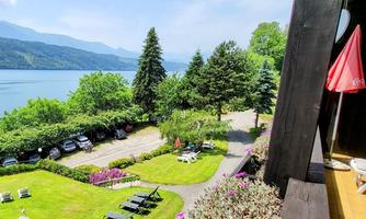 A picturesque view of a calm lake, surrounded by green trees and mountains. The garden is beautifully arranged with loungers and blooming plants.