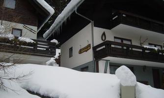 Two traditional mountain huts, surrounded by thick snow. The roofs are covered with snow drifts and there is a wintry atmosphere.