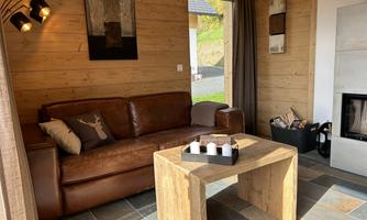 A cozy living room with a brown leather sofa and a wooden table. Large windows let in plenty of light and the walls are made of wood.