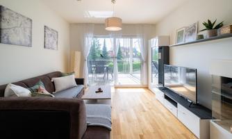 A modern living room with a cozy sofa and large windows. The space has a bright wooden floor and leads to a balcony with a view of the greenery.