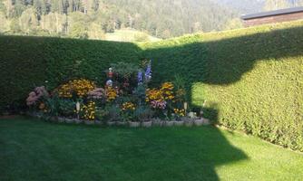 A well-kept garden with colorful flowers and a high privacy hedge made of bushes. In the background, gentle hills and trees are visible.