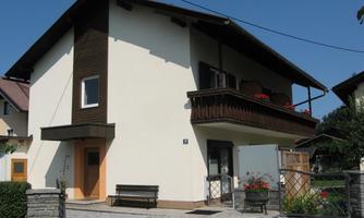 A two-story residential building with a balcony and light-colored walls. The entrance is surrounded by a small garden with flowers.