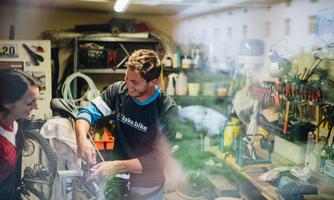 A bicycle mechanic is repairing a bicycle in his workshop. In the background, many tools and workshop utensils are visible.
