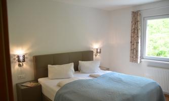 A cozy bedroom with a large double bed and soft lighting. The windows offer a view of greenery.