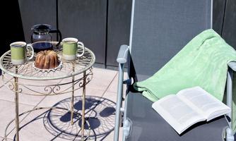 A cozy outdoor area with a small table, two cups, and a piece of cake. Next to the table lies an open book and a green blanket on a lounge chair.
