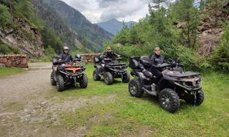 Three men on quads are standing on a narrow path in a green mountainous landscape. In the background, mountains and wooded areas can be seen.