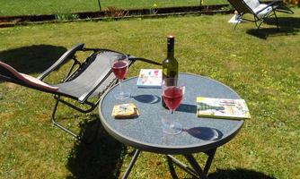 A small table with two glasses and a bottle of wine is set on a meadow. In the background, there are lounge chairs, and it is a sunny, relaxed atmosphere.