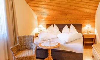 A cozy bedroom with a double bed and wooden paneling. Next to the bed is a small table with a chair.
