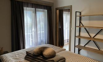 A modern bedroom with a large bed and striped bedding. There are large windows with curtains and an open wooden shelf.