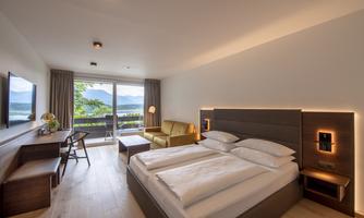 A modern hotel room with a double bed, a cozy armchair, and a desk. The large windows offer a beautiful view of the mountains, and the light creates an inviting atmosphere.