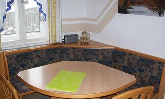 A cozy dining area with a round wooden table and upholstered benches. In the background, there are windows with curtains and a decorative bottle rack.