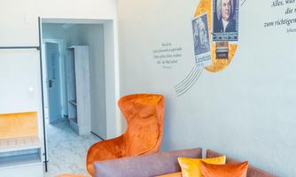A modern living room with a cozy sofa corner and orange decorative cushions. On the wall, there are pictures and inspiring quotes.