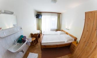 A simple bedroom with two single beds, a sink, and a small table. Large windows provide plenty of daylight.