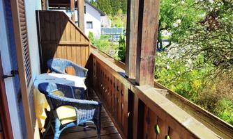 A balcony with a wooden railing and two chairs is ready for relaxing. In the background, green trees and a blue sky can be seen.