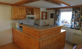 A modern kitchen with wooden details and a kitchen island. Daylight comes through a window and colorful curtains can be seen.