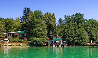 A tranquil lake with clear, green water and surrounding trees. In the background, colorful houses and a pier can be seen.