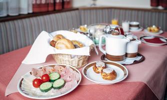 A laid breakfast table with various rolls, sausages, cheese, cucumbers, and tomatoes. Teapots and small plates with pastries are also visible.