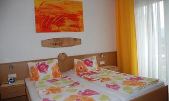 A cozy bedroom with two beds decorated with colorful pillows. The room has large windows with yellow curtains and a colorful painting on the wall.