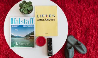 A round table with a magazine, a book, a plant, and a remote control. House slippers are on a red carpet.