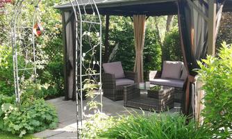 A cozy garden pavilion with two seating areas. Surrounded by green grass and vibrant vegetation.