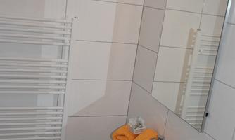 A modern bathroom with white tiles, a washing machine, and a towel holder. Yellow towels are on the floor, and a mirror is mounted on the wall.