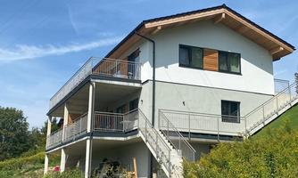 A modern house on a hill, surrounded by green spaces. The building has several balconies and large windows.