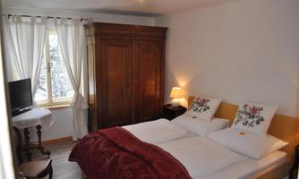A cozy bedroom with a double bed and a warm, inviting atmosphere. There is a wooden wardrobe and a window with bright daylight.