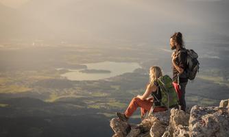 Two hikers are sitting on a rock, enjoying the view of a picturesque landscape with a lake. The sun is setting, bathing the scene in beautiful light.