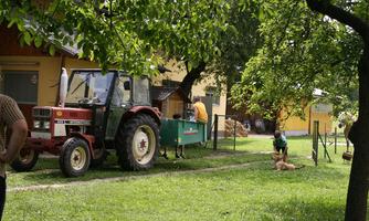 A red tractor is standing on a green meadow next to a house. In the background, two people are working while a dog is lying on the ground.