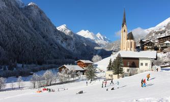 A picturesque winter village with snow-covered mountains and a church in the background. People are enjoying various winter activities in the snow.