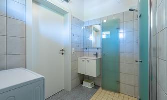 A modern bathroom with tiles and a glass shower. There is a wash area and a washing machine in the corner.