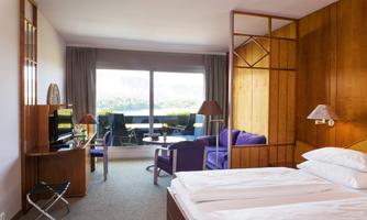 A modern hotel room with a double bed and warm wood details. The room offers a view of a balcony with a purple armchair and a beautiful view.