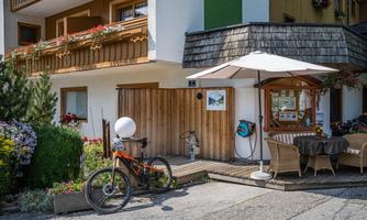 A cozy outdoor area with a table and chairs under a sunshade. Next to it stands a bicycle, and blooming plants adorn the entrance.