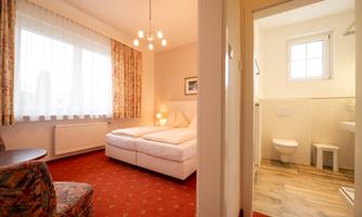 A cozy room with a double bed and a bright window. To the right is the bathroom with toilet and shower.