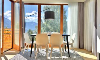 A bright dining area with a round table and white chairs. Large windows provide a view of the mountains and let in plenty of sunlight.
