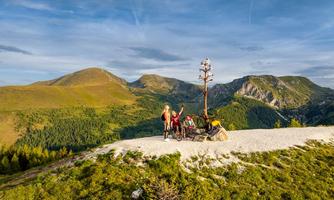 A group of people is standing on a green hill overlooking the mountains. In the background, steep cliffs and a clear sky can be seen.