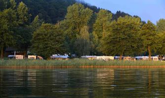 A quiet campsite by the shore of a lake, surrounded by tall trees. The water reflects nature and the caravan facilities.