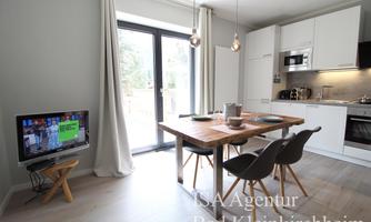 A bright, modern living room with a dining table and chairs. In the background is a kitchen and a television is visible.