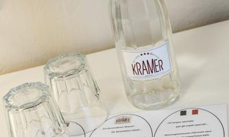 A glass bottle with the print "KRAMER" stands next to two overturned glasses on a table. Underneath lies a menu with various dishes.
