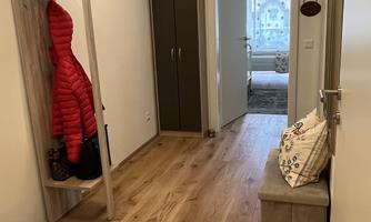 An inviting hallway with wooden flooring and warm lighting. A red jacket and a pair of shoes hang on the wall, while a door leads to another room.