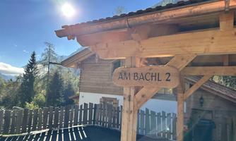A wooden house with a sign that says "AM BACHL 2". In the background, there are trees and a clear sky.
