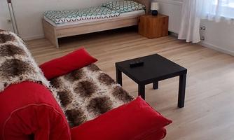 A cozy living room with a red sofa and a small black table. In the background, there is a bed and a piece of art featuring a mountain landscape.