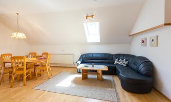 A cozy living area with a large black couch and a wooden table. The room has bright walls and plenty of daylight through a skylight.
